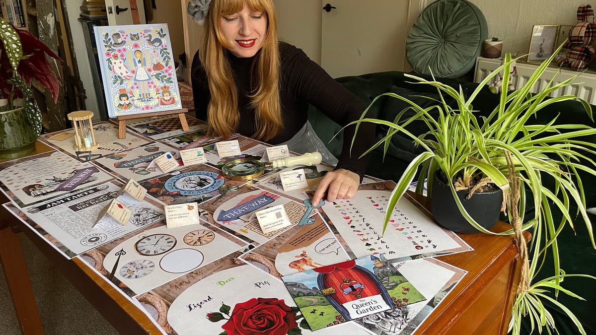 A woman sits behind a wooden table covered with printed cards, stickers, and small art pieces in a cozy indoor room. A potted green plant is prominent on the right, and an easel with a colorful illustration stands on the left.